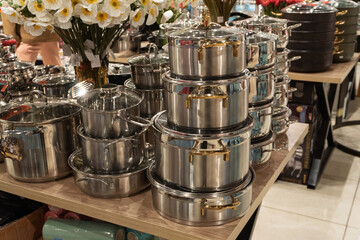 A variety of stainless steel pots and pans are neatly stacked on a wooden table in a retail store. The display includes different sizes of cookware, surrounded by artificial.