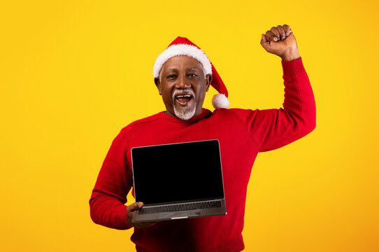 Senior black man wears a Santa hat and red sweater while happily showing a laptop with a website mockup. He raises his fist in excitement at a successful online Christmas sale event.