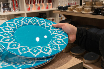A person holds a large blue plate with a white geometric pattern in a store. Shelves in the background display various dishes and kitchenware items.