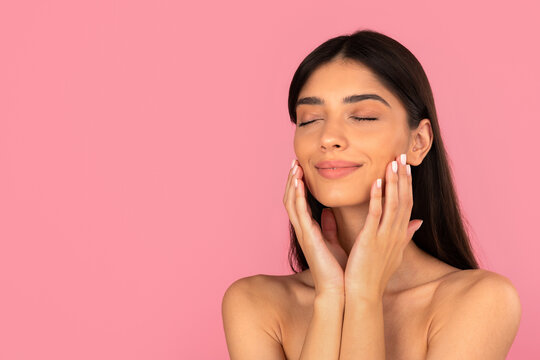 Relaxed young woman touching her face with eyes closed, enjoying skincare routine. Studio portrait showing calmness, self care and natural beauty concept