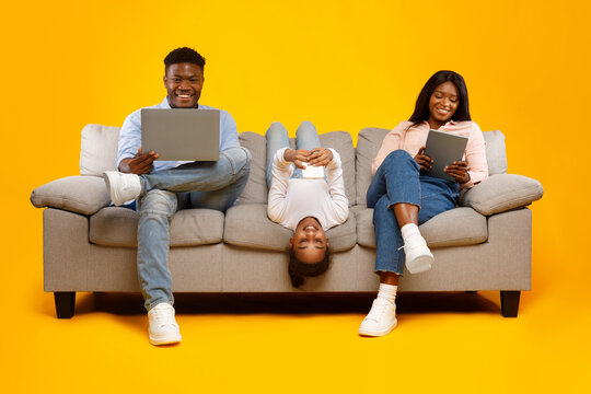 A joyful African American family of three engages with their electronic devices while sitting comfortably on a sofa. The warm yellow orange background enhances their bonding time.