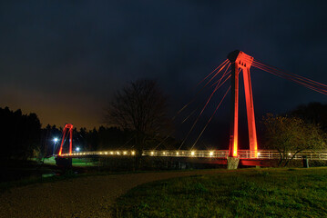 Night view of an illuminated pedestrian suspension bridge with red lights, glowing against a dark cloudy sky and forest background.