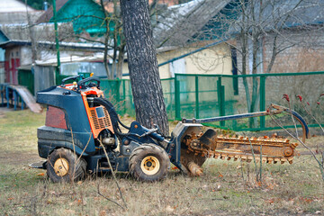 Walk-behind trencher parked in rural yard near tree prepared for digging, soil maintenance work in countryside environment. Small mechanical trenching machine resting in backyard, garden maintenance