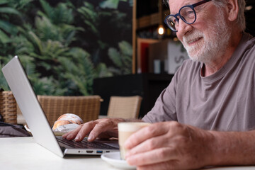 Smiling senior man sits in a cafe having break with coffee and croissant while using his laptop. An elderly business man uses technology expressing confidence and contentment in a relaxed atmosphere.