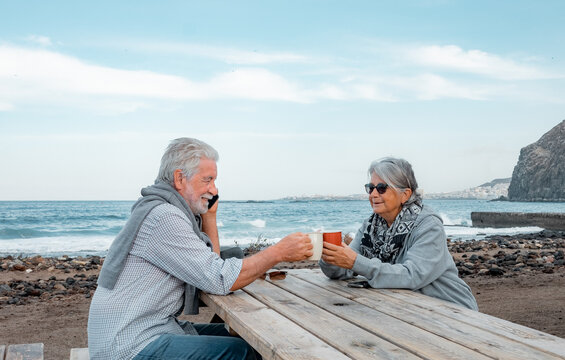 Smilling senior couple sitting for a break with a cup of tea along the seacoast while using smartphone. Serene retirement lifestyle - Powered by Adobe