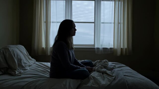 Sad woman sitting alone on her bed in dim light looking out the window in silence
