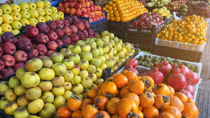 Fresh colorful fruits neatly arranged at outdoor market stand, showcasing apples, pomegranates, persimmons, oranges and other produce, symbolizing healthy nutrition, natural food and organic farming