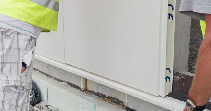 Close-up view of workers carefully aligning and lowering an insulated sandwich panel into position on a building wall.