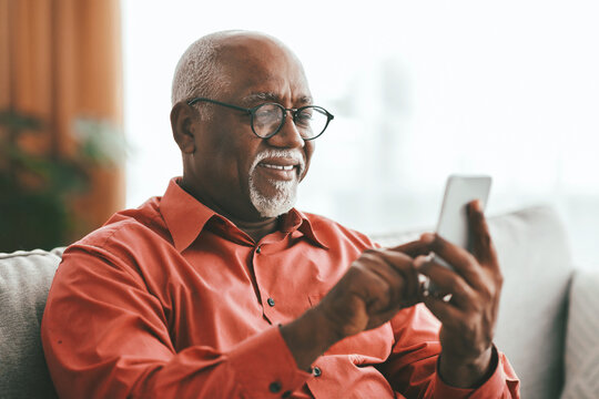 A smiling elderly man with glasses is interacting with his smartphone while sitting on a sofa. The bright, natural light from the window adds warmth to the cozy room. - Powered by Adobe