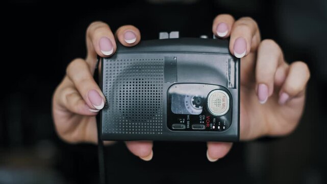 Front view of manicured hands holding a black portable cassette player with tape reels turning inside. This retro audio device represents 90s and 2000s music, nostalgia, and Y2K fashion.