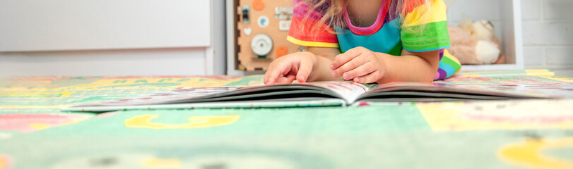 Toddler girl in bright clothes playing and learning with a book on a colorful floor mat at home.