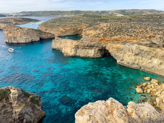 A spectacular panoramic view from a high vantage point over the rugged coastline and multiple coves of Comino Island, Malta. The clear turquoise waters are navigated by a tourist cruise boat