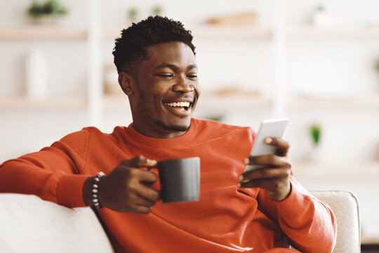 A young man with short curly hair sits comfortably on a sofa, holding a cup and looking at his smartphone with a big smile. The bright room is filled with greenery and soft decor.