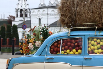 A car with hay on the roof, with a full interior of yellow and burgundy apples against the background of a white church