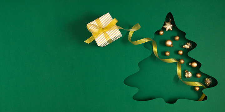 Green Christmas background with a cutout in the shape of a Christmas tree decorated with golden baubles and a star. Framed with a gold ribbon and a gift. The bright winter background has space for tex - Powered by Adobe