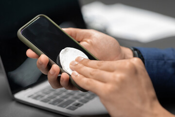 A businessman wipes his smartphone with sanitizer while seated at a desk. This closeup shows the importance of cleanliness during the pandemic in a busy office setting.