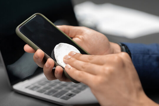 A businessman wipes his smartphone with sanitizer while seated at a desk. This closeup shows the importance of cleanliness during the pandemic in a busy office setting. - Powered by Adobe