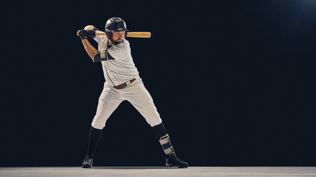 Baseball player in full uniform holding bat in batting stance on dark background. Concept of professional sport preparation, performance focus, and athletic training marketing.