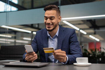 A cheerful entrepreneur from the Middle East sits at his desk in an office. He is using his smartphone while holding a credit card, enjoying online shopping and managing his finances.