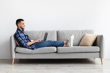 Obraz premium Young Arab man sits comfortably on a sofa in his living room, smiling while browsing the internet on his laptop. Casual setting with a relaxed atmosphere for personal enjoyment.