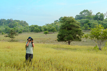 Naklejka premium Asian cameraman photographer standing holding camera in the middle of a meadow. Male tourist shooting grass field and tree landscape. Backpacker adventure taking photo