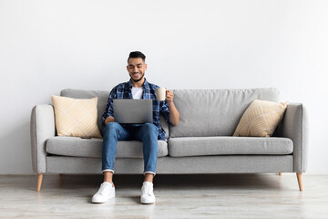 A cheerful Arab man is seated on a cozy couch, watching a video on his laptop while sipping morning coffee. He appears relaxed and engaged, enjoying his time at home.