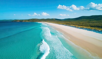 An aerial view of a pristine white sandy beach with turquoise ocean waves and green hills in the distance