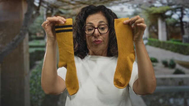 Middle aged hispanic woman holds mismatched socks with both hands while looking at them in park; playfulness individuality spontaneity.