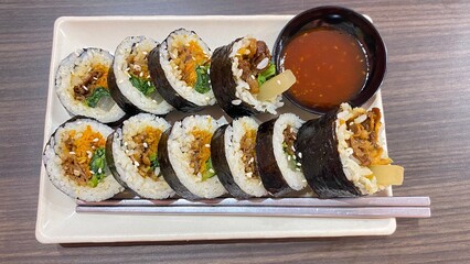 Korean kimbap is served on a white plate with chopsticks and sauce in a small bowl. The vegetables, meat, and rice are wrapped in fresh seaweed.