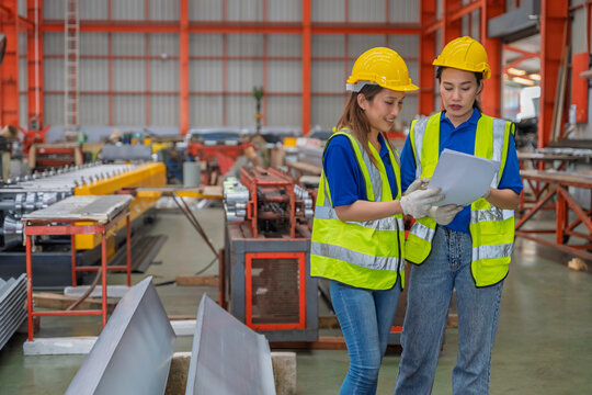 Two female factory workers wearing safety helmets and reflective vests operating industrial machine in manufacturing plant. Women engineers in industry workplace. - Powered by Adobe