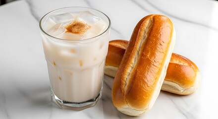 Iced creamy beverage with cinnamon topping next to two golden brown bread rolls on a marble surface