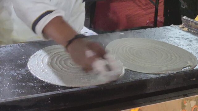 A chef prepares a fresh dosa on a hot plate, with visible steam and golden texture forming on the flat surface.