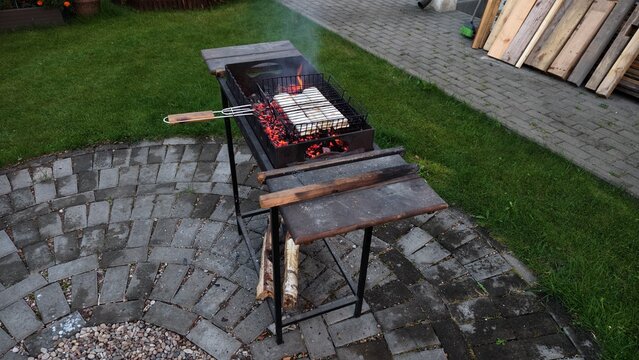 There is a metal brazier on the tiled area, in which coals are smoldering and lavash with cheese is being prepared on the grill. There are firewood nearby. Next to it is a grassy lawn Evening twilight - Powered by Adobe