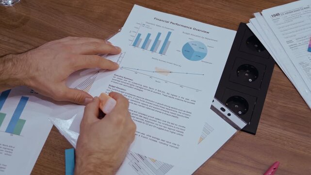 Detailed view of a business employee marking key points on a financial performance overview, emphasizing graphs and numerical data on a wooden table with scattered papers and visible outlets