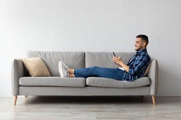 Portrait shows a young Arab man comfortably sitting on a grey sofa at home. He is using his smartphone to send messages while enjoying his free time in a cozy living room setting.