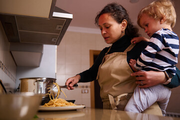 Mother Cooks Pasta With Child On Hip In Kitchen, Wearing Apron, Focused On Meal Preparation