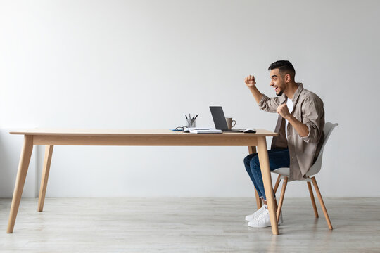 An Arab man is sitting at a desk in a home office, celebrating success. He is excitedly shaking his fists and shouting yes while looking at his laptop, clearly thrilled with the news he just received.