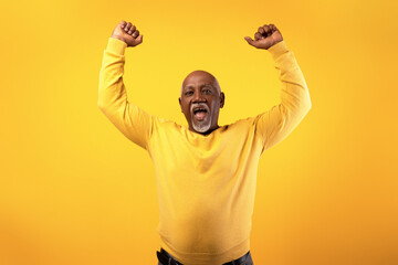 Fototapeta premium An overjoyed senior black man expresses excitement by shouting and raising his arms in triumph. He is set against a bright orange background, showcasing his happiness and enthusiasm for success.