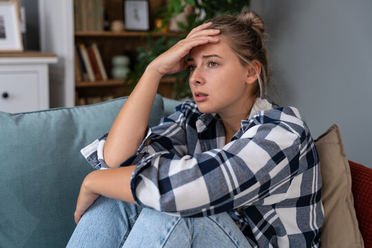 Young woman at home struggling with depression and anxiety, holding her head on sofa; signs of burnout, loneliness and overthinking, seeking health support and calm after stress, and sleepless nights