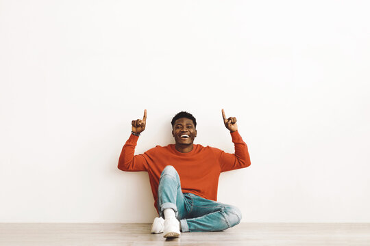 A young man sits on a wooden floor, wearing a red sweater and blue jeans. He raises his fingers in excitement, expressing joy in a brightly lit indoor space.
