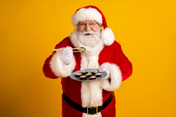 Santa Claus in a red costume with white fur hat and beard offers sushi on a plate to celebrate Christmas against a bright yellow background