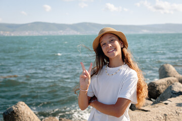 Smiling girl in a hat standing by the sea on a sunny day, showing a peace sign and enjoying summer vacation. Concept of freedom, happiness, travel, and connection with nature.
