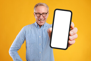 Excited adult man wearing glasses holds a white smartphone close to the camera. He smiles in a vibrant yellow-orange studio backdrop, promoting a website with a blank phone display.