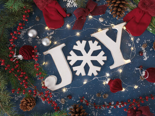 Overhead Christmas background with JOY letters, red gloves mittens, festive velvet burgundy ornaments and bows, holiday twinkle lights and red berries, evergreen branches, pinecones on blue backdrop