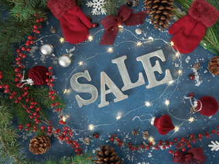 Overhead Christmas background with SALE letters, red gloves mittens, festive velvet burgundy ornaments and bows, holiday twinkle lights and red berries, evergreen branches, pinecones on blue backdrop