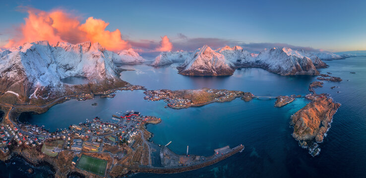 Aerial panoramic view of Reine village and surrounding fjords in Lofoten Islands, Norway. Snow-covered mountains, blue sea, colorful houses illuminated by soft sunrise light in winter. Top drone view - Powered by Adobe