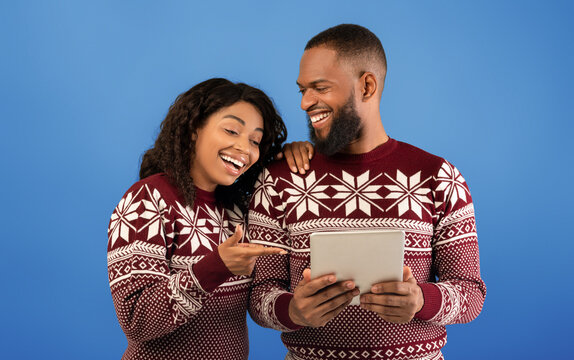 Excited black couple in warm sweaters using digital tablet over blue background, doing online Christmas shopping together, standing over blue background, copy space