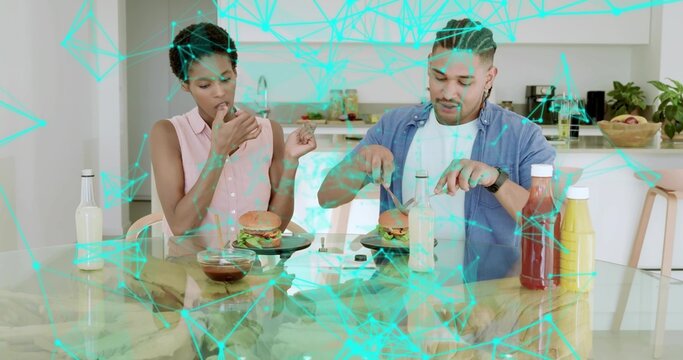 Cutting burger with knife and fork, couple wearing pink top, denim at kitchen table, turquoise grid - Powered by Adobe