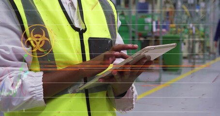 Holding tablet, worker wearing yellow vest tapping screen in factory aisle, with biohazard overlay