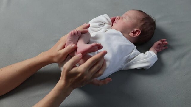 A newborn baby in a white bodysuit against a gray background. The little one is sleeping. The child is resting with its legs spread out. The mother holds the baby's tiny feet in her hands.